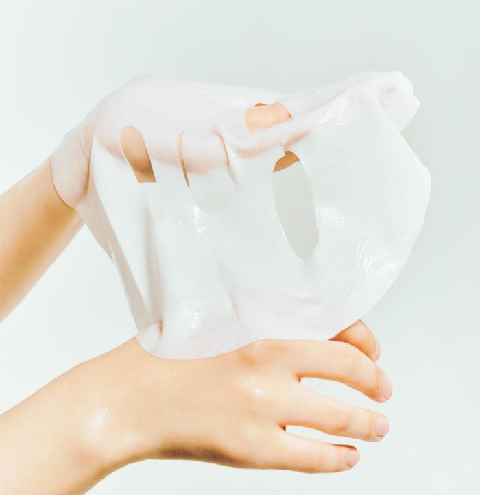 Hand holding a face sheet mask against a light gray background